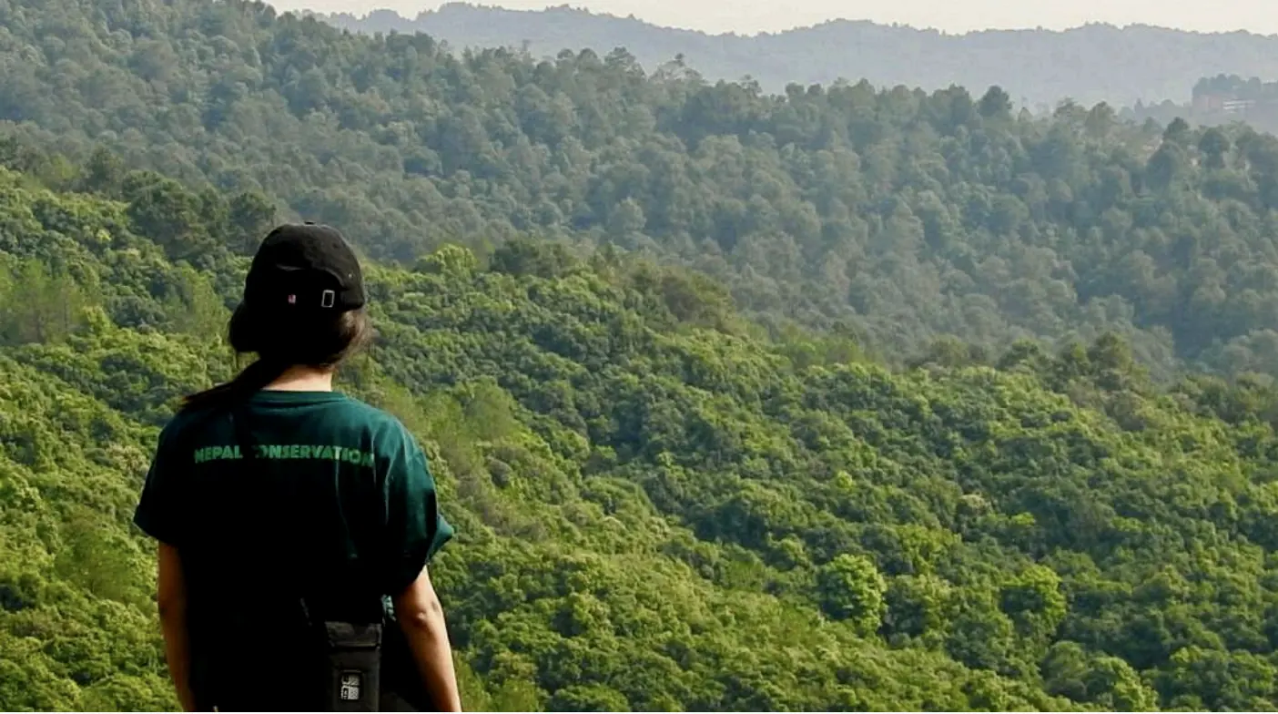 A student in a Nepal Conservation shirt looking out over forested ridges